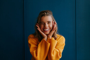 Young blond hair woman in yellow sweater touching face and winking while standing on blue background