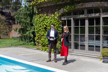 Fototapeta na wymiar Realtor and potential client observe big swimming pool near veranda in suburban cottage. Real estate manager and businesswoman examine offer