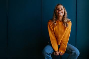 Beautiful young blond hair woman in yellow sweater smiling while sitting on blue background