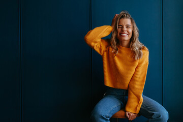 Playful young blond hair woman holding hand in hair and winking while sitting on blue background - obrazy, fototapety, plakaty