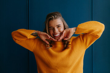 Young blond hair woman in yellow sweater touching face and winking while standing on blue background