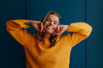 Young blond hair woman in yellow sweater touching face and smiling while standing on blue background