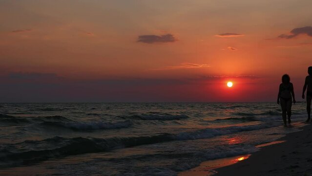 A man and a woman are walking along the seashore in front of a beautiful sunset. Slow Motion