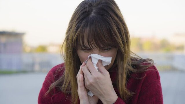 An attractive middle-aged Caucasian woman woman blows her nose into a handkerchief and coughs - busy road in the blurry background