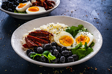 Boiled eggs with boiled white rice, sun dried tomatoes and green olives on wooden table
