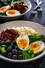 Boiled eggs with boiled white rice, sun dried tomatoes and green olives on wooden table
