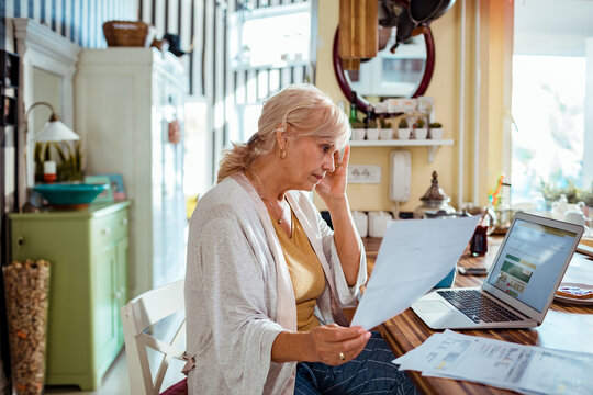 Stressed senior woman holding bill using laptop at home
