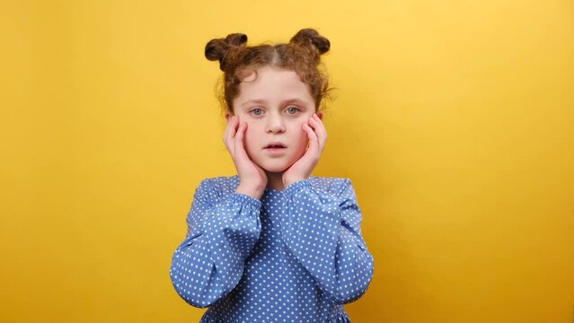 Omg. Portrait of surprised little girl child opening mouth and touching face, looking at camera in shock, posing isolated over plain yellow color background wall in studio. People emotion concept