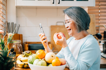 A grandmother, an old woman, is having breakfast in the kitchen. She's smiling, using her smartphone, and eating a red apple. A modern portrait of technology and happiness at home. - obrazy, fototapety, plakaty