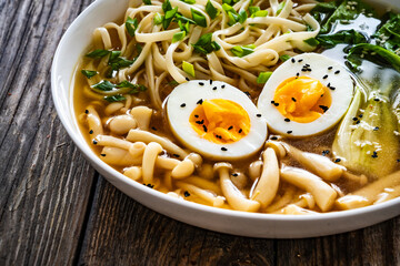 Asian style soup. Broth with chow mein noodles, boiled eggs, shimeji mushrooms and pak choi on wooden table
