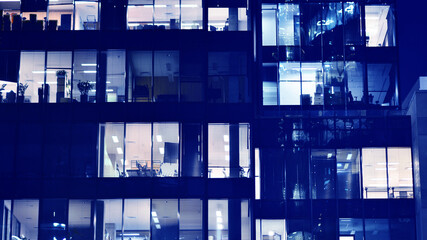 Fragment of the glass facade of a modern corporate building at night. Big glowing windows in modern office buildings at night, in rows of windows light shines. Blue graphic filter.