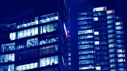 Fragment of the glass facade of a modern corporate building at night. Big glowing windows in modern office buildings at night, in rows of windows light shines. Blue graphic filter.