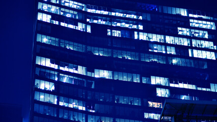 Fragment of the glass facade of a modern corporate building at night. Big glowing windows in modern office buildings at night, in rows of windows light shines. Blue graphic filter.