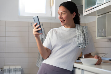 A housewife girl stands in the kitchen with a phone and a plate in her hands and blogs about...