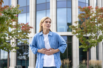 Mature woman in a shirt is walking in the city, a worker has a severe stomach ache, a blonde businesswoman in casual clothes is sick, massaging her hands with her hands outside an office building. - obrazy, fototapety, plakaty