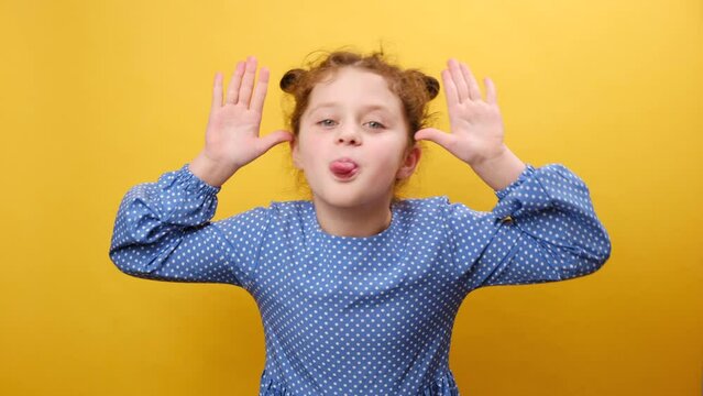 Portrait of funny little girl child sticking out tongue and grimacing looking at camera, holding hands near temples, posing isolated over plain yellow background wall in studio. Misbehavior concept