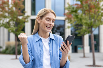 Fototapeta premium Happy successful winner woman in blue casual shirt received online notification message on phone, mature business woman outside office building celebrating triumph, joyfully holding hand up.