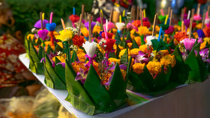A photo of several bright green banana leaf krathongs with petals carefully arranged on a clean white future board. Inside the krathong are bright yellow marigolds, purple orchids, artificial flowers