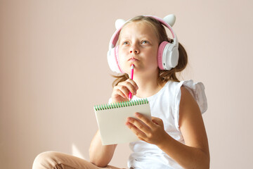 An 11-year-old blonde girl holds a notebook in her hands, headphones on her head, looking thoughtfully to the side