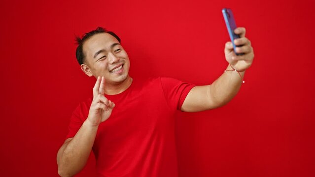 Young chinese man smiling confident making selfie by the smartphone over isolated red background