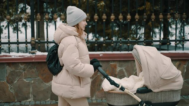 
Woman in winter gear walking with a baby stroller along a snowy park path, with ornate metal fence and bare trees in the background.
