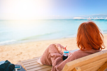 Red-haired girl from behind is lying on chaise longue covered with a blanket. Woman is resting on the sea beach and admiring the ocean landscape. Alcoholic drink in female's hands. Summer holidays.