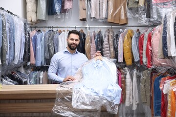 Dry-cleaning service. Happy worker holding hangers with clothes in plastic bags at counter indoors