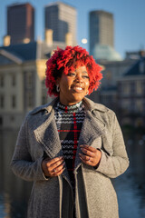 Happy smiling female model with strong unique style appearance in urban inner city environment The Hague cityscape skyline. Confident black lady stylish red hair with winter good taste Holland fashion