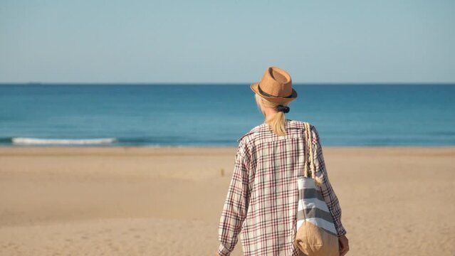 Beautiful young woman in hat and beach shirt walking on vast sandy beach with bag, heading to sea on tall dunes. Place without people Patara Beach in Turkey.