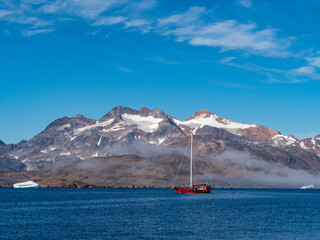 Ein rotes Segelschiff ohne Segel fährt vor schneebedeckten Bergen in den Fjord von Tasiilaq.