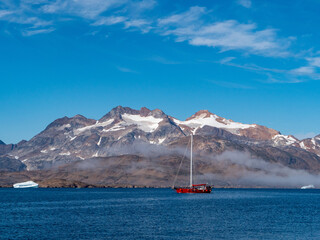 Ein rotes Segelschiff ohne Segel fährt vor schneebedeckten Bergen in den Fjord von Tasiilaq.
