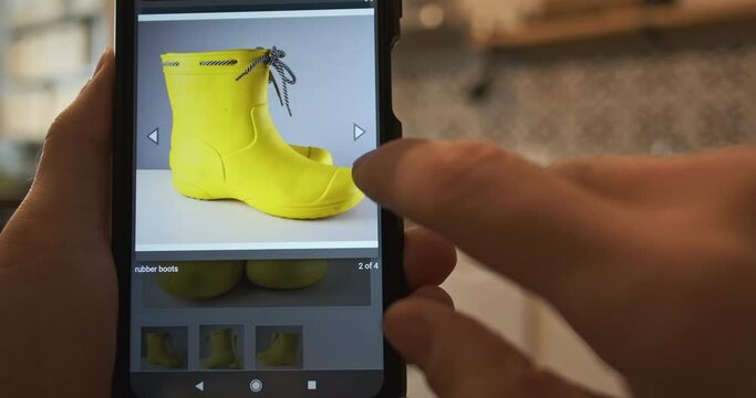 Examine pictures of shoes on smartphone screen. The concept of online shopping, choose a product, flip through the photos of yellow rubber boots on the website of an online shopping mall.