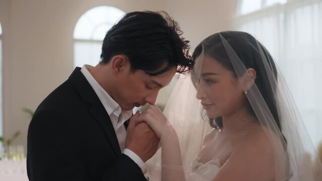 Romantic Asian groom gently kissing hand of beautiful bride in veil during wedding ceremony.