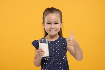 Cute girl with glass of fresh milk showing thumb up on orange background