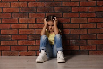 Child abuse. Upset little girl sitting on floor near brick wall indoors