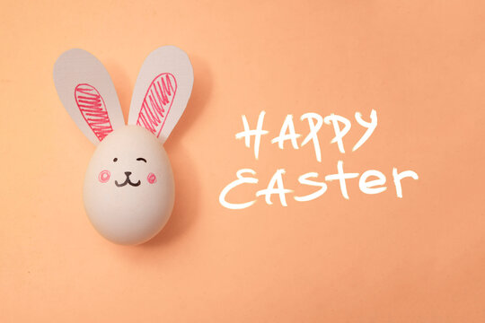 Food photo. Chicken egg with cute bunny face and rabbit ears on beige background. Postcard for family holiday. Text Happy Easter. Preparation for celebration. Trendy pastel peach color of 2024. 