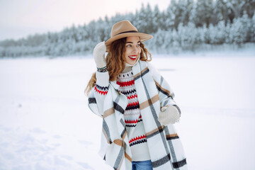 Fototapeta premium Portrait of a smiling woman in a hat and scarf in a snowy forest enjoying a winter day. Young traveler posing outdoors. Adventure, weekend concept.