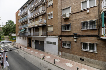 A city street with four-story residential buildings made of dark bricks and leading to a park with...