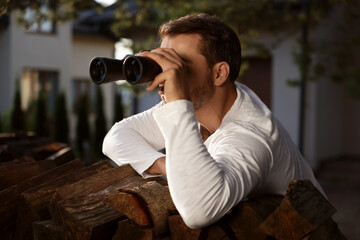 Fototapeta na wymiar Concept of private life. Curious man with binoculars spying on neighbours over firewood outdoors