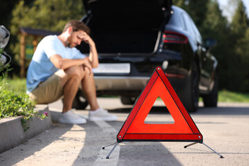 Man sitting near broken car on roadside outdoors, focus on warning triangle