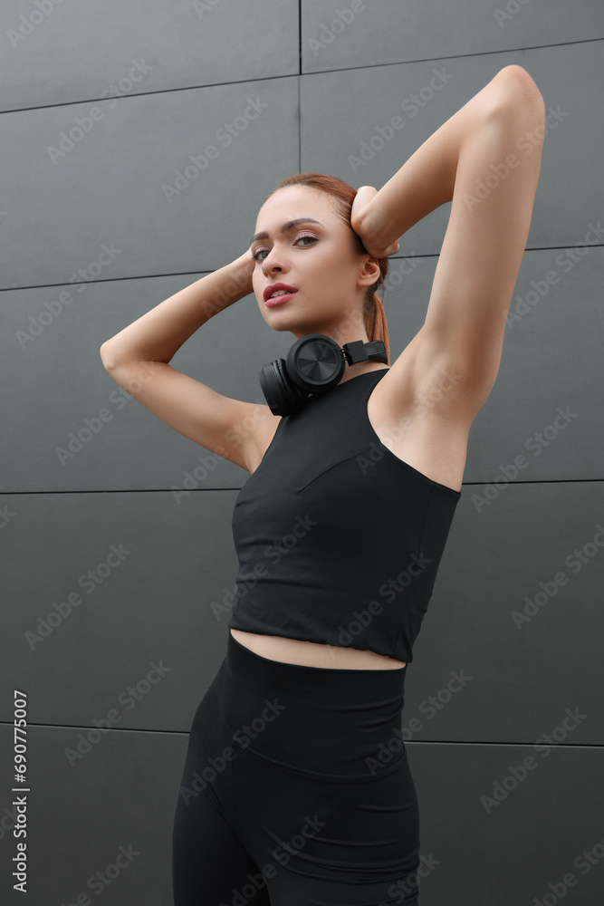 Poster Beautiful woman in stylish gym clothes with headphones posing near dark grey wall on street