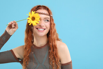 Beautiful young hippie woman covering eye with sunflower on light blue background, space for text