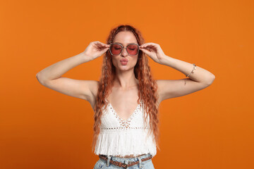 Stylish young hippie woman in sunglasses sending air kiss on orange background