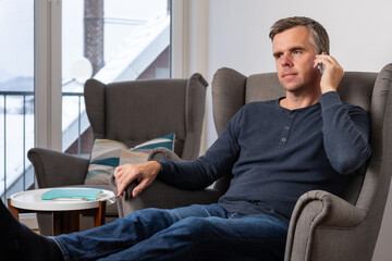 handsome 40s single  man sits in an comfy  grey armchair and listens to a call on his smartphone
