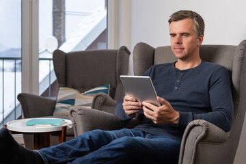 handsome 40s single  man sits in an comfy  grey armchair and reads on tablet

