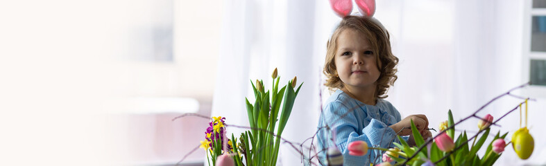 Portrait of cute pretty smiling little girl in blue dress and bunny rabbit ears preparing easter table decor. Fresh spring bright colourful flowers in the kitchen. Family time. Banner copy space