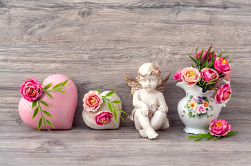 Valentine's day. The angel statue is decorated with a decorative heart and flowers in a vase. A gift for lovers. Beige wooden background.