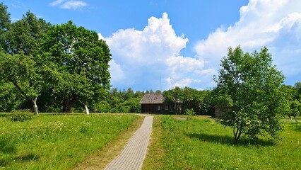 An old wooden house with a tiled roof and lightning rod sits next to an orchard with whitewashed trunks. A tiled path leads to the house through a grassy lawn. Sunny summer weather and blue skies