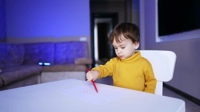 Thoughtful kid sits at desk holding a red pen. Toddler drawing a picture sitting at desk.