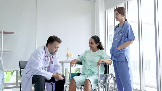 Middle age male doctor communicating and having check right leg of caucasian patient women in wheelchair at clinic. Client has positive for cure result. Nurse standing to support.